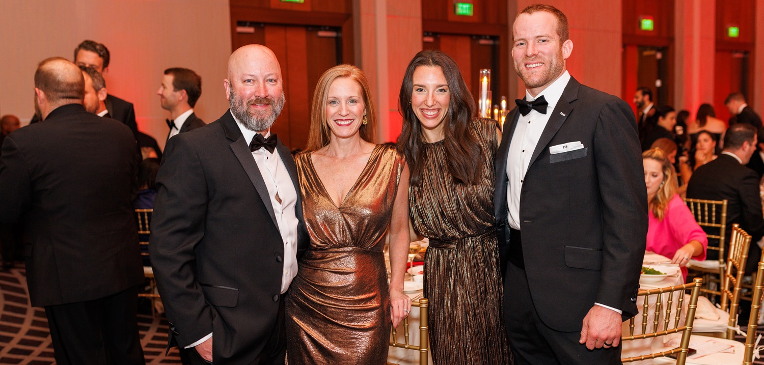 Two couples in formal wear standing together in a crowded ballroom.